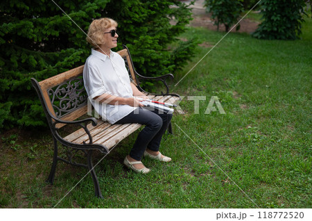 An elderly blind woman sits on a bench in the park with a folded tactile cane in her hands. An elderly blind woman sits on a bench in the park with a folded tactile cane in her hands. 118772520