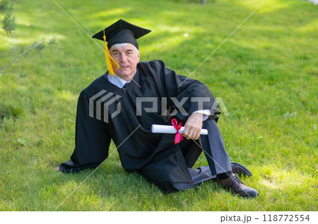 An elderly gray-haired man in a graduation gown sits on green grass. An elderly gray-haired man in a graduation gown sits on green grass. 118772554