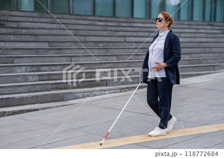 Blind pregnant businesswoman walking along tactile tiles with a cane. Blind pregnant businesswoman walking along tactile tiles with a cane. 118772684