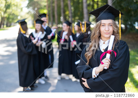 Group of happy students in graduation gowns outdoors. A young girl with a diploma in her hands in the foreground. 118772860