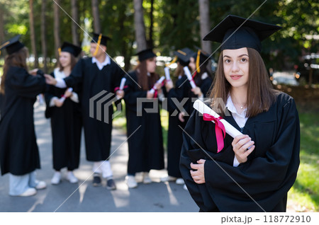 Group of happy students in graduation gowns outdoors. A young girl boasts of her diploma. Group of happy students in graduation gowns outdoors. A young girl boasts of her diploma. 118772910