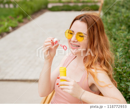 Young red-haired woman blowing soap bubbles outdoors. Girl with braces on her teeth.  118772933