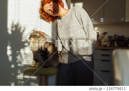portrait red-haired curly young woman with pet gray fluffy cat in arms, lit by bright sun portrait red-haired curly young woman with pet gray fluffy cat in arms, lit by bright sun 118779811