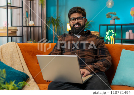 Happy young Indian man typing working on laptop browsing internet using apps sitting on home sofa 118781494