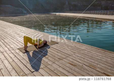 A Woman Engaging in Yoga Practices at the Serene Waterfront During the Beautiful Sunrise Hour 118781768