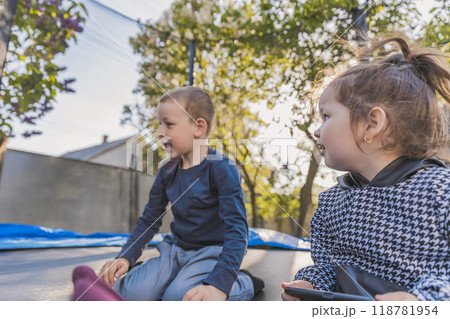 Two children sitting outdoors, smiling and engaging with each other in a playful environment, surrounded by trees and soft lighting Two children sitting outdoors, smiling and engaging with each other in a playful environment, surrounded by trees and soft lighting 118781954