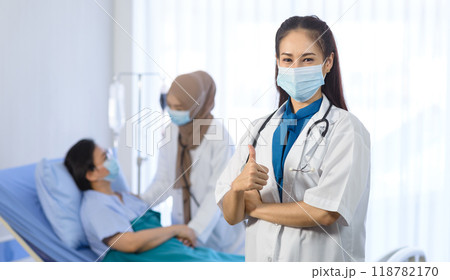 Young asian female doctor wear face mask arms crossed and showing thumbs up while standing in font of muslim doctor checking elderly patient on background. medical team working in the hospital room. Young asian female doctor wear face mask arms crossed and showing thumbs up while standing in font of muslim doctor checking elderly patient on background. medical team working in the hospital room. 118782170