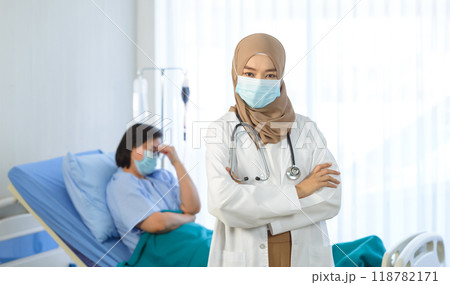 Young muslim female doctor arms crossed stand in hospital with elderly patient lie down on bed on the background. Young muslim female doctor arms crossed stand in hospital with elderly patient lie down on bed on the background. 118782171