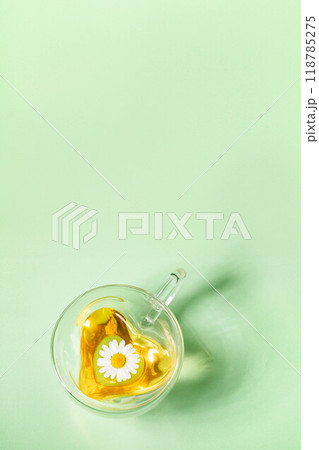 Heart glass cup with chamomile tea with one flower on light green vertical background. Copy space. Heart glass cup with chamomile tea with one flower on light green vertical background. Copy space. 118785275