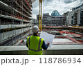 Engineer reviewing a large civil engineering project blueprint at a construction site. The architect wears overalls, a vest and a helmet. Engineer working at site of a large building project. 118786934
