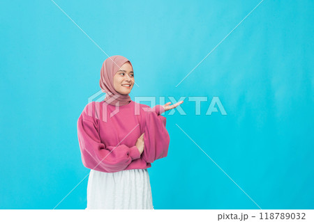 A joyful woman in a pink sweater and colorful hijab stands out against a vibrant blue background 118789032