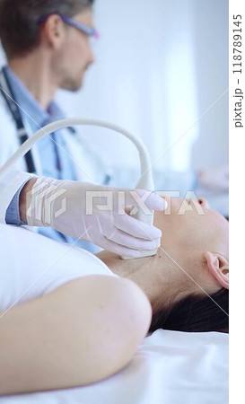 Doctor man wearing white protective gloves is using ultrasound machine to examine neck of female patient in hospital. Medicine, thyroid gland health Doctor man wearing white protective gloves is using ultrasound machine to examine neck of female patient in hospital. Medicine, thyroid gland health 118789145