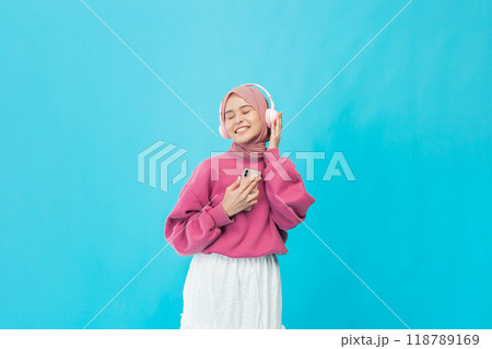 A carefree young woman enjoys music in a stylish pink sweater against a vibrant blue background 118789169