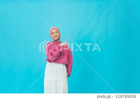 Cheerful Young Woman Dressed in a Bright Pink Sweater Pointing Joyfully Against a Blue Background Cheerful Young Woman Dressed in a Bright Pink Sweater Pointing Joyfully Against a Blue Background 118789202