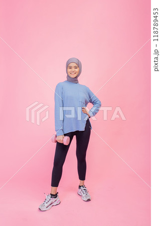 An athletic woman holds a dumbbell in stylish activewear against a vibrant pink background 118789453