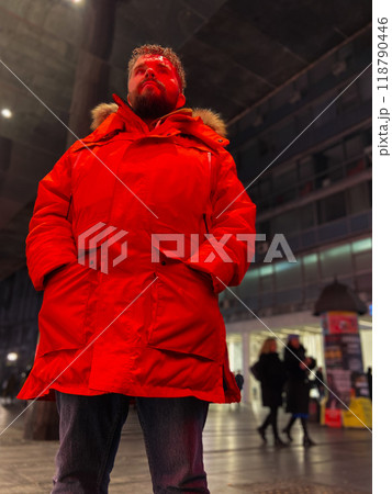 Man walking in a bustling nighttime city street, wearing a bright orange parka. Red neon light. Serene contemplation against the backdrop of lively city lights at night. Urban life concept 118790446