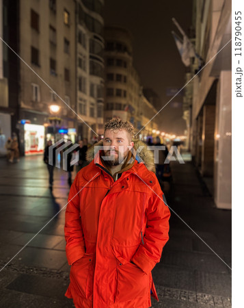 Man walking in a bustling nighttime city street, wearing a bright orange parka. Serene contemplation against the backdrop of lively city lights at night. Urban life concept 118790455