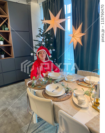 Millennial woman in a Santa suit sits thoughtfully and sad at a festive dining table, surrounded by Christmas decorations and a lit tree. Moment of holiday reflection and problem loneliness in 118790470