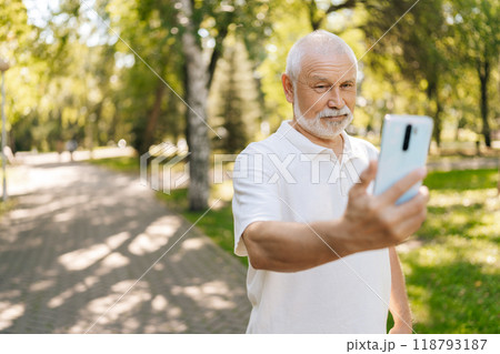 Portrait of handsome white-bearded senior man taking selfies on smartphone while walking on summer sunny park surrounded by lush greenery and trees. Happy elderly male capturing selfie in park. Portrait of handsome white-bearded senior man taking selfies on smartphone while walking on summer sunny park surrounded by lush greenery and trees. Happy elderly male capturing selfie in park. 118793187