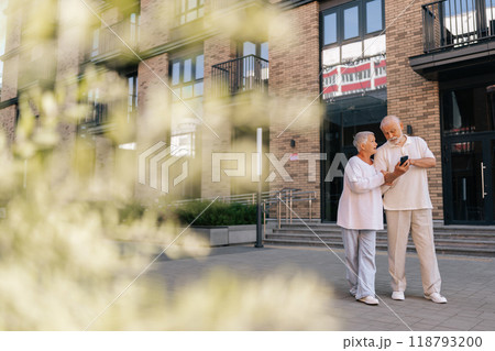 Wide remote shot of beautiful elderly couple exploring new city streets, relying on smartphone for directions. Gray-haired senior male and female navigating unfamiliar city with smartphone navigation. 118793200