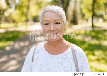 Portrait of beautiful gray-haired senior woman smiling looking at camera standing posing in green summer park on sunny day, exuding confidence and contentment in casual outfit, enjoying retirement 118793201