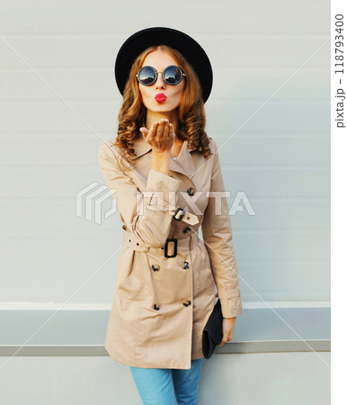 Fashion pretty young woman blowing kiss wearing a black hat sunglasses coat over grey background Fashion pretty young woman blowing kiss wearing a black hat sunglasses coat over grey background 118793400