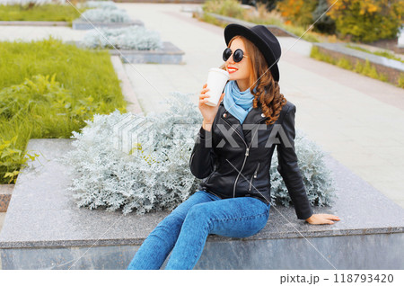 Fashion cool girl drinks coffee from cup wearing a black rock jacket, hat in city park Fashion cool girl drinks coffee from cup wearing a black rock jacket, hat in city park 118793420