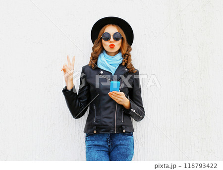 Fashion cool girl using phone blowing a kiss wearing a black rock jacket hat on white background 118793422