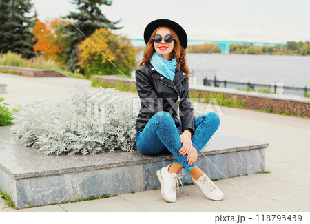 Fashion young smiling woman posing wearing black rock jacket, hat in city park 118793439