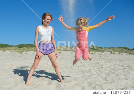 Two girls are playing on a sandy beach. One is jumping joyfully as sand flies up, while the other stands nearby, smiling. The clear blue sky and greenery create a peaceful summer backdrop Two girls are playing on a sandy beach. One is jumping joyfully as sand flies up, while the other stands nearby, smiling. The clear blue sky and greenery create a peaceful summer backdrop 118794287