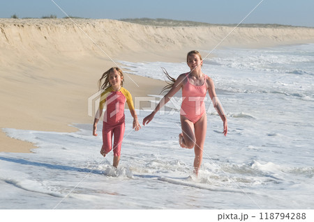 Two girls are running joyfully along the shoreline, splashing through the shallow waves on a sunny beach. The sandy dunes and clear sky create a perfect summer day atmosphere full of fun. Two girls are running joyfully along the shoreline, splashing through the shallow waves on a sunny beach. The sandy dunes and clear sky create a perfect summer day atmosphere full of fun. 118794288