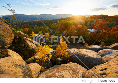 Setting sun casts golden glow over autumn landscape, illuminating vibrant red and orange foliage. Rugged rock formations in foreground under cloudy sky. Dovbush Rocks, Carpathian mountains, Ukraine. 118801553