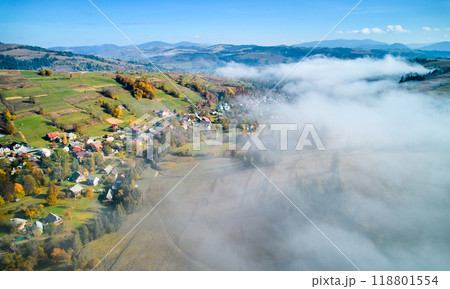 Picturesque village nestled in rolling hills, bathed in autumn colors with patches of green and yellow fields. Wisps of fog drift through landscape, adding dreamy atmosphere to serene rural scene. Picturesque village nestled in rolling hills, bathed in autumn colors with patches of green and yellow fields. Wisps of fog drift through landscape, adding dreamy atmosphere to serene rural scene. 118801554