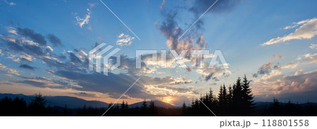 Sun sets behind range of distant mountains, casting golden rays through clouds. Sky, painted with shades of blue, pink, and gold, in tranquil evening. Silhouetted pine trees add depth to landscape. 118801558