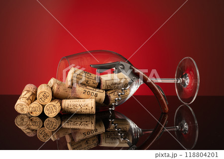 A wine glass filled with wine corks lies on a black glass plate and is reflected in it, in front of it stands a fine wine cutlery set with a wooden handle against a red background A wine glass filled with wine corks lies on a black glass plate and is reflected in it, in front of it stands a fine wine cutlery set with a wooden handle against a red background 118804201