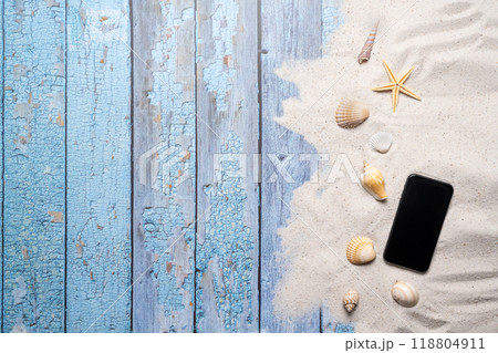 Summer, beach and vacation concept with free text space. Top view. Flat layout with a smartphone, various sea shells and fine beach sand on an old blue wooden boards background Summer, beach and vacation concept with free text space. Top view. Flat layout with a smartphone, various sea shells and fine beach sand on an old blue wooden boards background 118804911