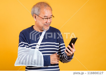 Portrait Asian old man cast his arm and holding mobile phone studio isolated yellow background, retired elderly man accident broken arm wear splint treatment, insurance medical care application Portrait Asian old man cast his arm and holding mobile phone studio isolated yellow background, retired elderly man accident broken arm wear splint treatment, insurance medical care application 118808127