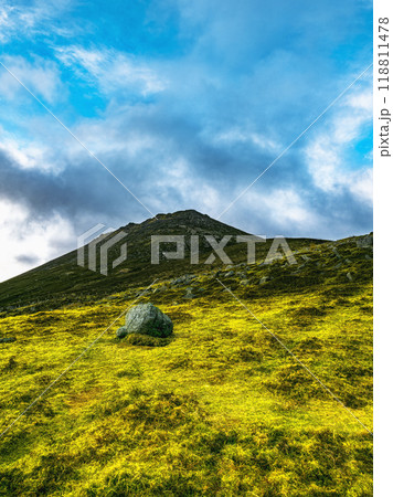 A large, rounded rock sits in the foreground of a grassy hill. The hill slopes upward towards a mountain summit under a partly cloudy sky. 118811478