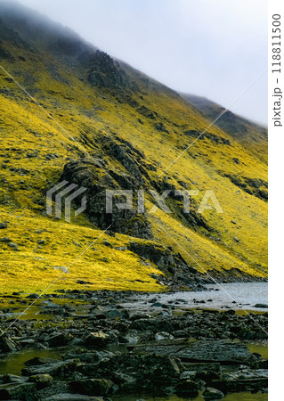 A rocky, grassy hillside slopes down towards a small river. The water is calm, and the riverbed is covered in dark rocks. The sky is a hazy white, suggesting overcast conditions. A rocky, grassy hillside slopes down towards a small river. The water is calm, and the riverbed is covered in dark rocks. The sky is a hazy white, suggesting overcast conditions. 118811500