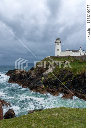 Storm clouds gather above Fanad Head Lighthouse as ocean waves crash against rocks in dramatic scene 118819075