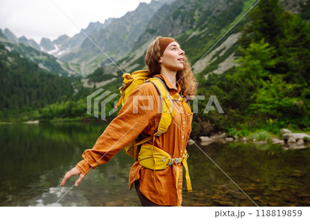 Female traveler with yellow hiking backpack against the backdrop of mountain lake with hiking poles. 118819859