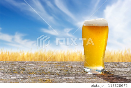 A glass of cold beer sits on a table with a vast rye field 118820673
