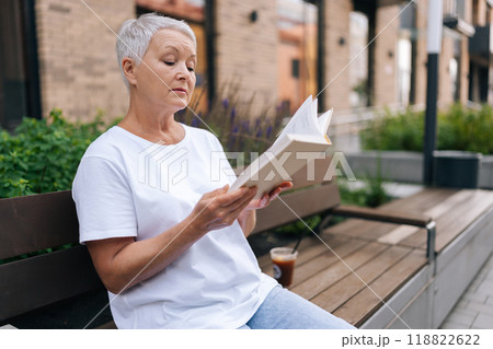 Focused elderly woman finding peace and tranquility reading book in urban street. European gray-haired older female enjoying fresh air while reading paper book in city environment. Focused elderly woman finding peace and tranquility reading book in urban street. European gray-haired older female enjoying fresh air while reading paper book in city environment. 118822622