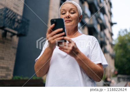 Low-angle view of gray-haired senior lady using smartphone texting message standing on urban street on summertime. Middle aged woman grandma hold phone typing sms enjoying communication in mobile app. Low-angle view of gray-haired senior lady using smartphone texting message standing on urban street on summertime. Middle aged woman grandma hold phone typing sms enjoying communication in mobile app. 118822624