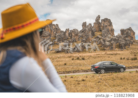 A woman wearing a yellow hat is looking at a car driving down a road, traveling through the peruvian andes. 118823908