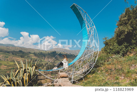 A girl sits on a blue hammock in a field with mountains in the background, traveling through the peruvian andes. Lima, Peru. 118823969