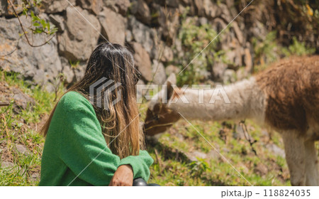 A woman sits on the ground next to a brown and white llama, traveling through the peruvian andes. Canta. Lima, Peru. 118824035