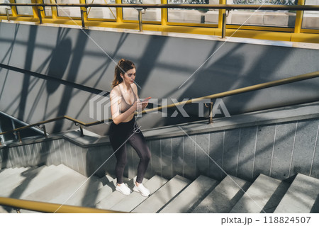 Mid-20s Caucasian woman taking a break on the stairs of a modern pedestrian bridge, checking her smartphone with earphones. Wearing a sports bra and leggings Mid-20s Caucasian woman taking a break on the stairs of a modern pedestrian bridge, checking her smartphone with earphones. Wearing a sports bra and leggings 118824507