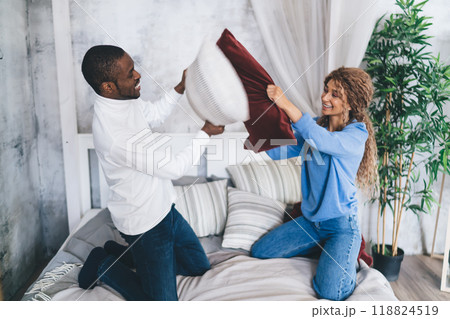 Joyful interracial couple engaging in a playful pillow fight in a bright, modern bedroom 118824519
