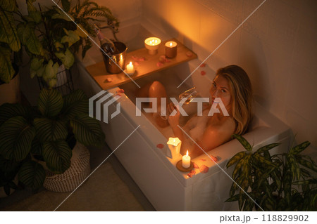 A young beautiful woman lies in a bath with foam and rose petals by candlelight. A young beautiful woman lies in a bath with foam and rose petals by candlelight. 118829902